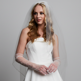 A woman wearing a white bridal gown and pearl and rhinestone gloves, standing against a green leafy backdrop.